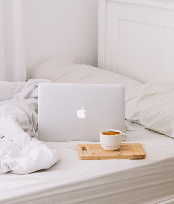 silver laptop next to a wooden serving tray with a white mug full of coffee on white luxury bedding