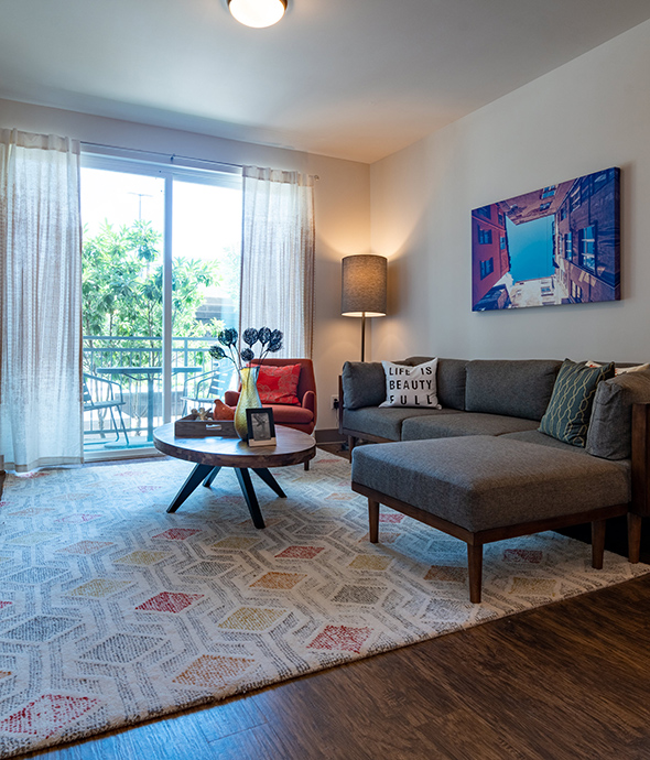 Living room with rich wood floors with rug, comfortable sectional couch, round coffee table, and sliding door to balcony.
