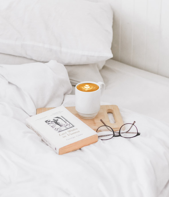 detail of white luxury bed with a book and reading glasses on a wooden tray with a white mug filled with coffee on bed