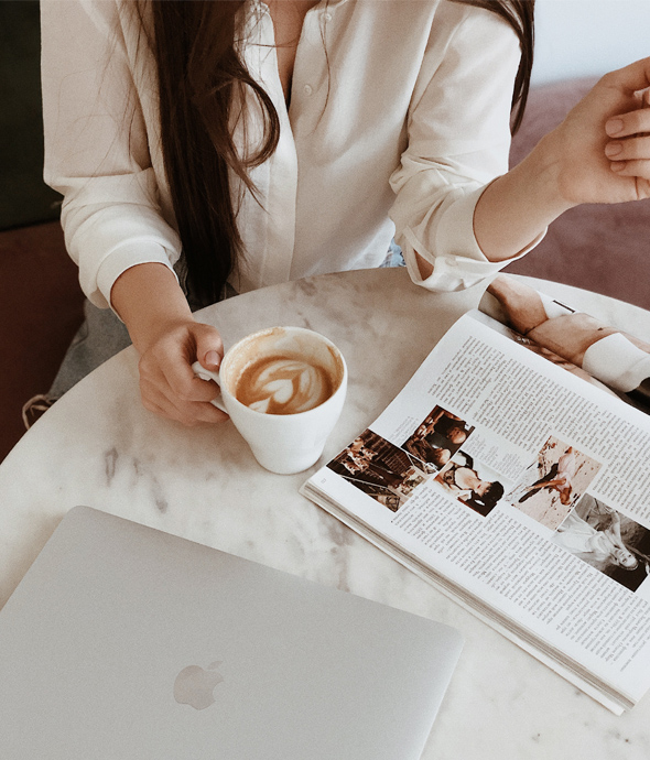 person sitting at a marble with table holding a cappuccino near laptop and reading a magazine