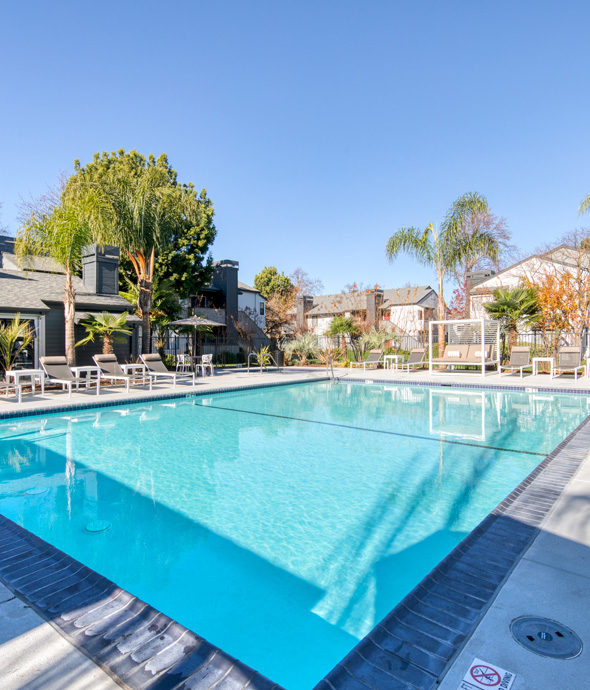 outdoor pool area surrounded by sitting chairs and tables with luxury loungers all shaded by beige umbrellas and area connected to Pleasanton clubhouse