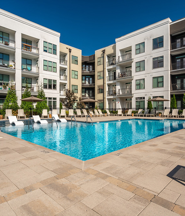 Pool area with stone pavers, bright blue water, and lounge chairs with umbrellas overlooked by apartments.