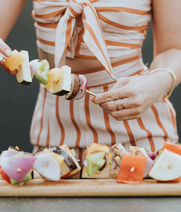 Person in dress prepares skewer with large pieces of vegetables.