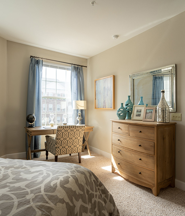 Bedroom with floral bedspread, tall wood dresser with bottles on top, plush carpet, and desk in front of tall window.