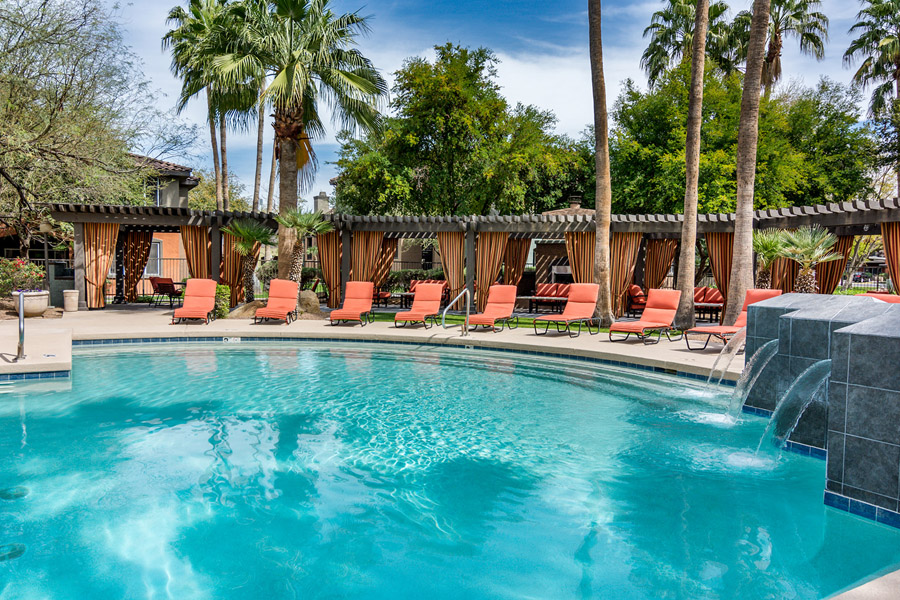 Mesa pool area with lounge chairs, tall palm trees, and long pergola with fireplace encircling deep blue pool.
