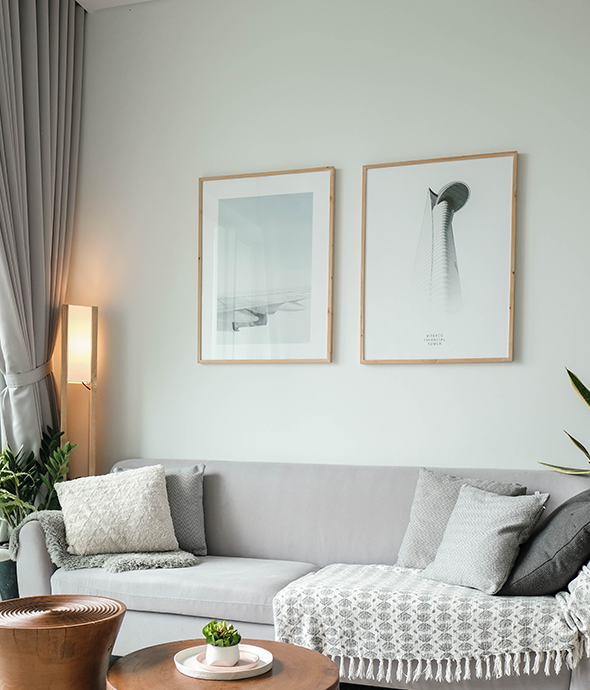 A grey sofa with pillows, artwork hanging above and a lamp next to window