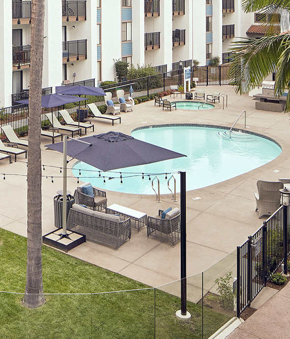 View from balcony of pool area with outdoor furniture, umbrellas, and tall palm trees.